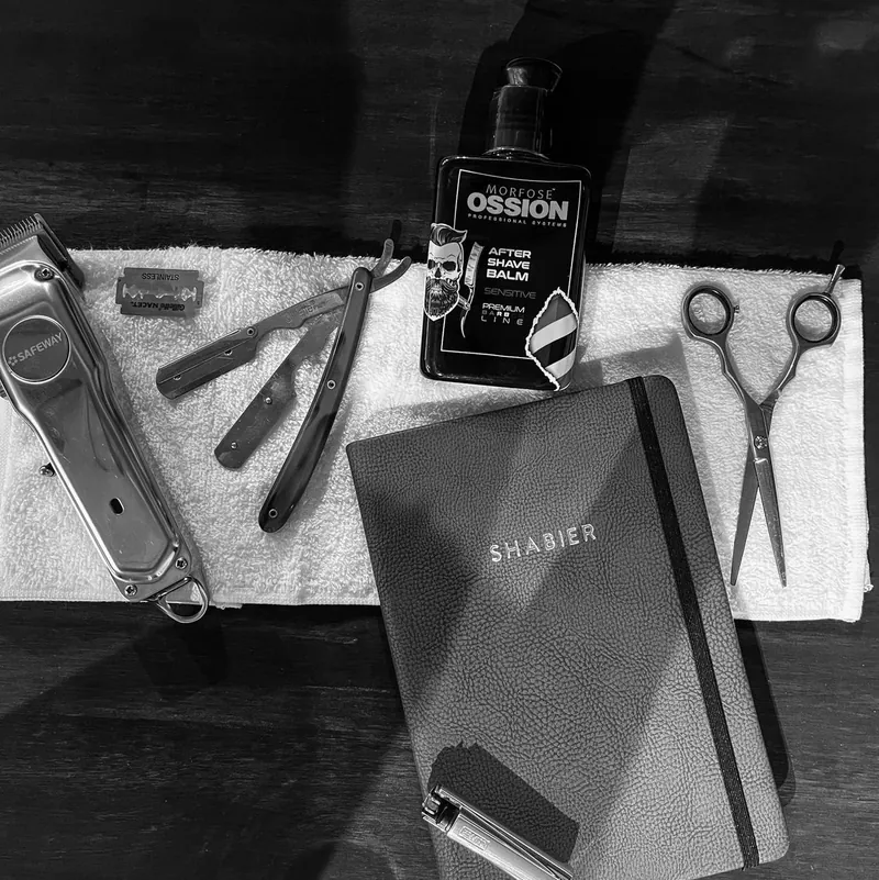 Shabier's tools laid out on a towel: clippers, straight razor, scissors, after-shave balm and a notebook embossed SHABIER